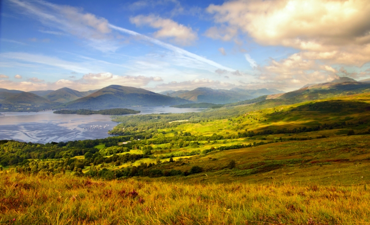 Hügelige Landschaft mit See und bewölktem Himmel im Hintergrund.