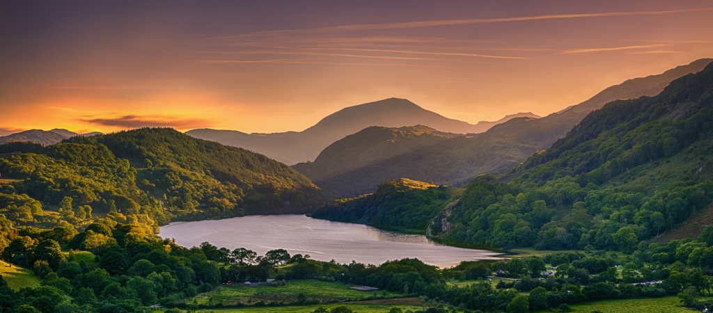 Panoramablick auf eine grüne, bergige Landschaft bei Sonnenuntergang mit einem See im Vordergrund.