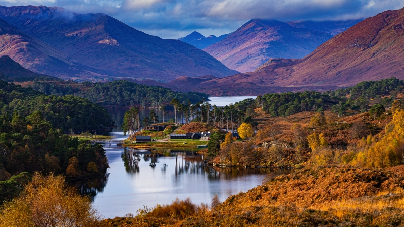 Hügelige Landschaft mit See, Bäumen und bewölktem Himmel.