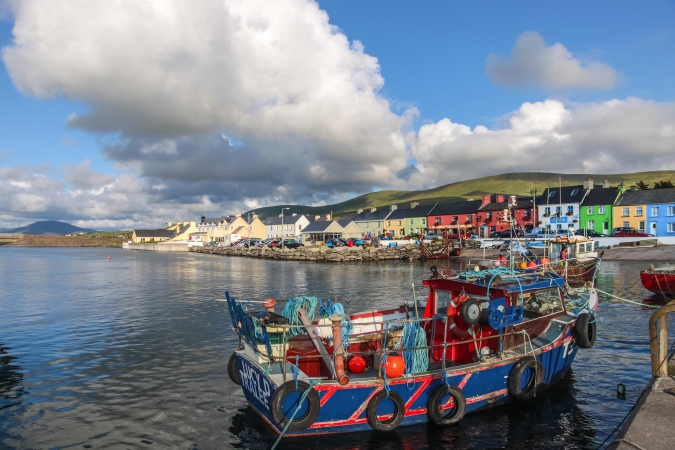 Fischerboote im Hafen von Portmagee, mit bunten Häusern im Hintergrund und bewölktem Himmel.