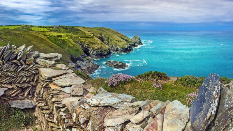 Küstenlandschaft mit Felsen und blauen Meer, im Vordergrund eine Steinmauer.